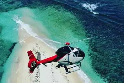 Helicopter passing above a small sandy islet, with shallow turquoise water and darker ocean water beyond.